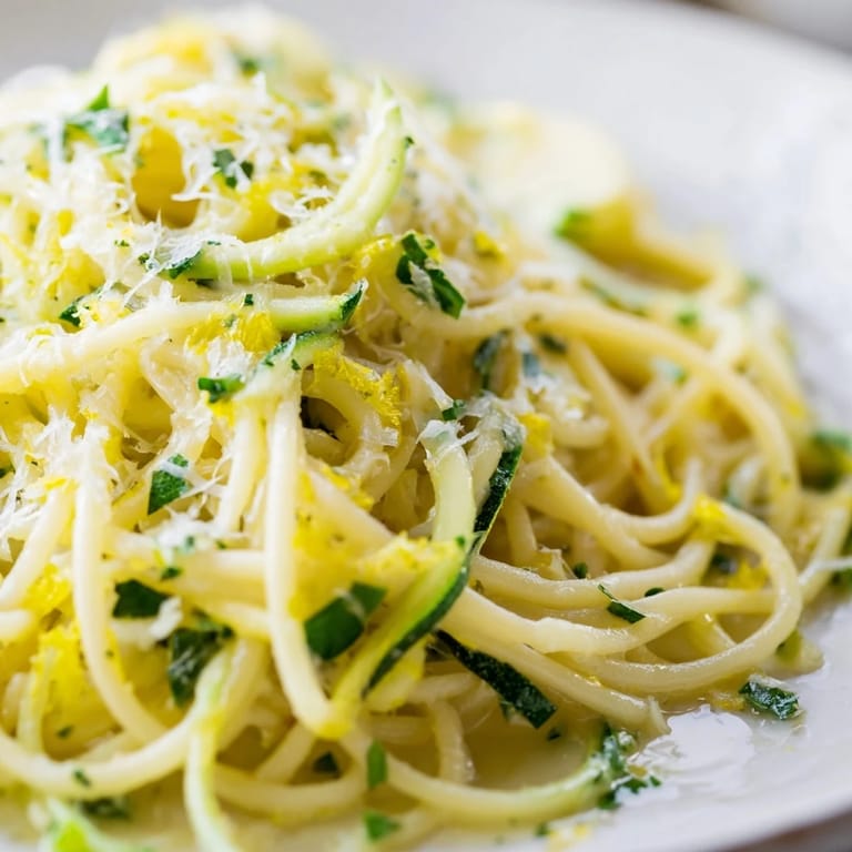 An overhead view of Lemon Zucchini Pasta served in a white bowl, highlighting the vibrant green zucchini spirals and a sprinkle of black pepper and red pepper flakes beside a glass of white wine.